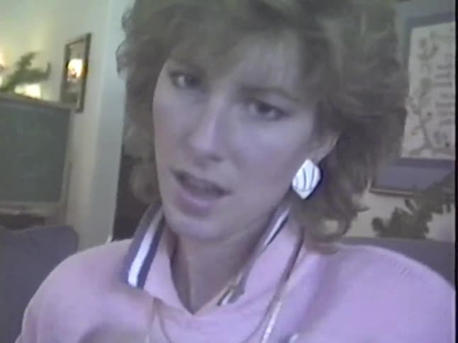 A young woman in the 1980s makes a silly face at the camera, stretching her mouth wide before breaking into laughter in a vintage home video