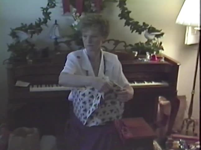 A woman carefully unwraps a Christmas gift while sitting in front of a piano in this nostalgic home video from the 1980s