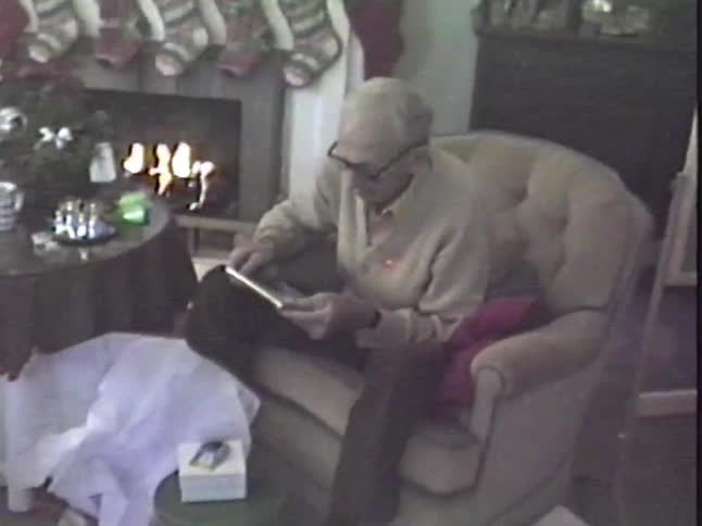 An elderly man opens a Christmas present while sitting in an armchair next to a warm, cozy fireplace in a vintage home video