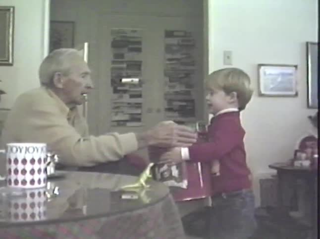 A family gathers on Christmas morning as a toddler excitedly opens a large gift tin with his grandfather in this vintage home video