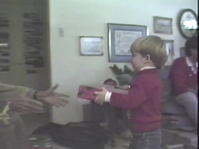 Vintage home video shows a young boy handing a Christmas present to his grandfather during a family holiday gathering