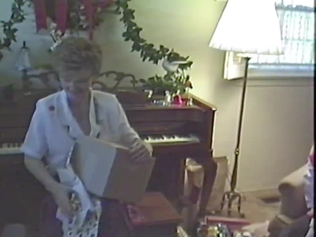A woman smiles as she carefully opens a Christmas present in a cozy living room in this retro home movie