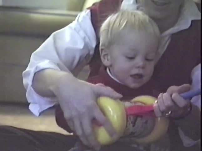 A cute baby boy sits on his mom's lap and opens a colorful new toy for Christmas in this vintage home video