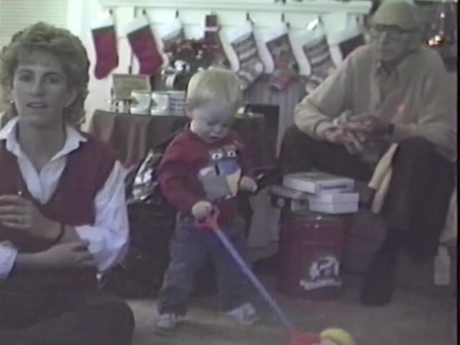 A toddler boy plays with his new push toy on Christmas morning as his proud mother and grandfather watch in a vintage home movie