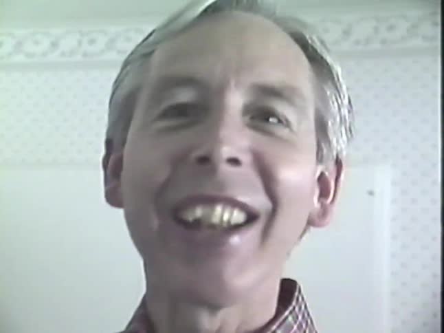 An older man wearing a plaid shirt fiddles with an old stove, then turns to the camera and smiles