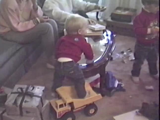 A vintage home video shows a toddler standing in a large toy dump truck in a living room surrounded by Christmas presents