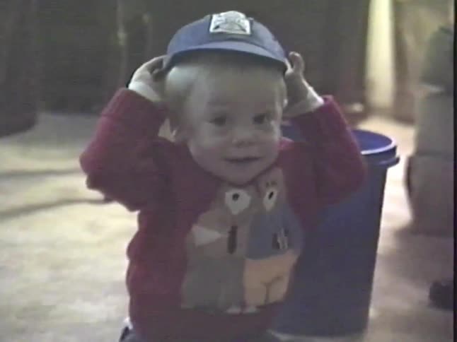 An adorable blonde baby boy sits on the floor and plays with his baseball cap in vintage home video footage