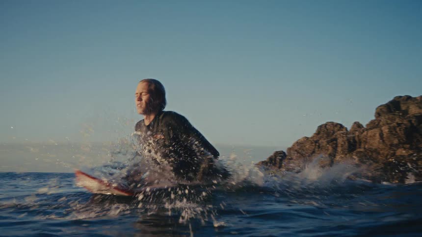 In slow motion, a focused surfer leaps from coastal rocks onto his board to paddle out into the ocean