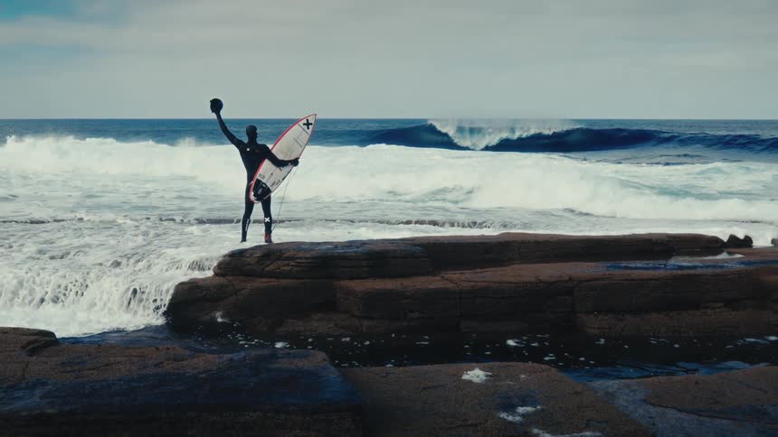 Cinematic slow motion of a surfer holding his board and celebrating on a rocky shoreline against a backdrop of powerful, curling ocean waves