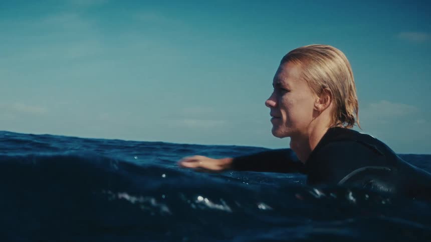 A young athletic surfer with blonde hair paddles out in the open ocean in slow motion