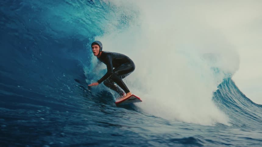 A skilled surfer in a full wetsuit masterfully rides inside the barrel of a massive blue ocean wave in dramatic slow motion