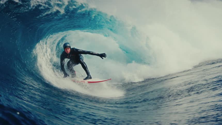 A surfer expertly rides inside the barrel of a massive, powerful ocean wave in dramatic slow motion