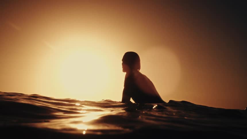 A silhouetted surfer waits patiently on their board, floating on gentle waves as the golden sun sets over the ocean