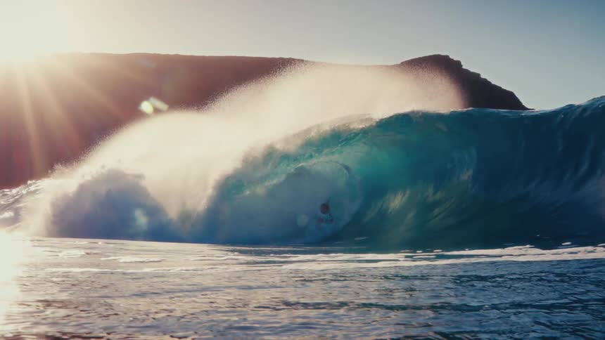 An expert surfer carves through the inside of a massive, beautiful turquoise wave in slow motion during a golden sunset