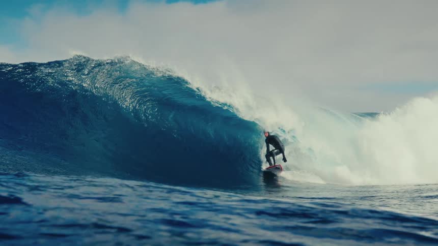 An expert surfer in a full wetsuit masterfully carves inside a massive blue ocean wave in breathtaking slow motion