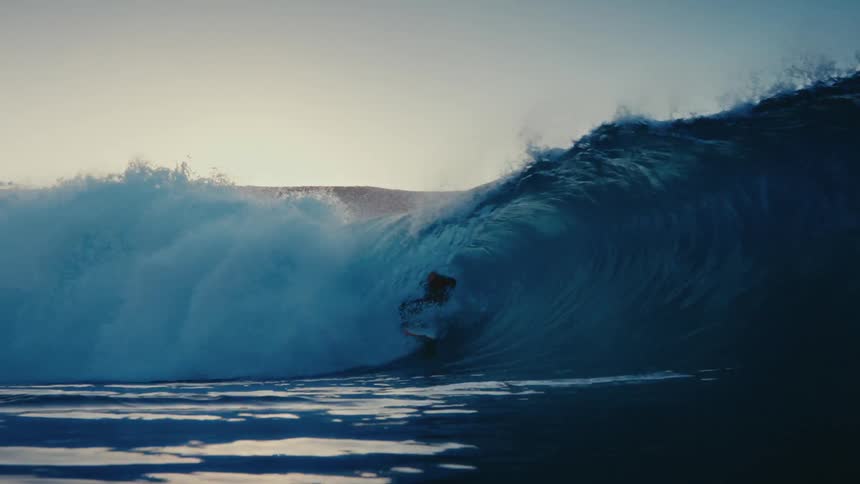 A surfer expertly rides inside the barrel of a powerful ocean wave in slow motion at sunset