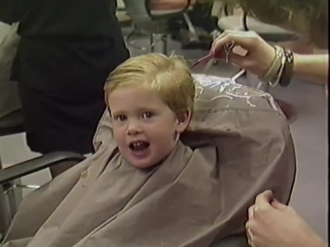 A cute toddler makes funny faces, going from surprised to happy, while getting his hair cut at the salon