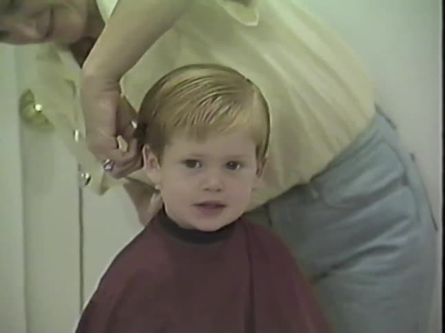 A vintage home video shows a happy little boy smiling while his grandmother gives him a haircut in their home