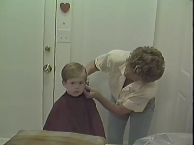 Vintage home movie footage shows a woman giving a young boy a haircut at home with scissors and a comb