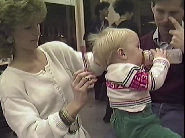 Vintage VHS footage shows a stylist giving a young boy his first haircut as his dad holds him and he drinks from a bottle