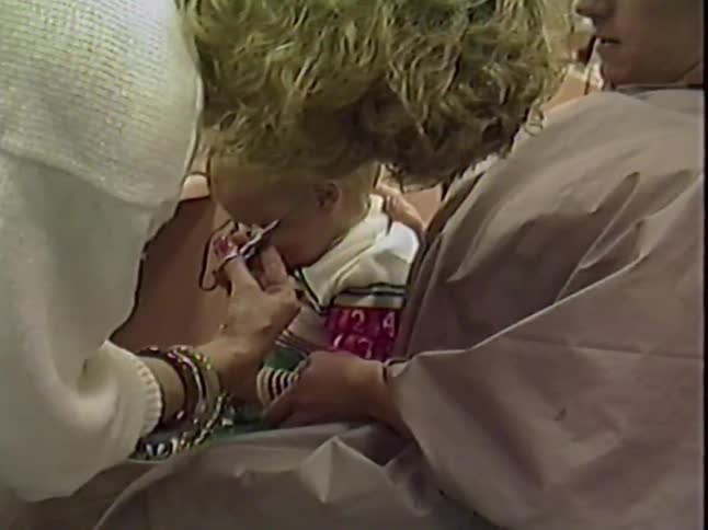 A young blond toddler gets his very first haircut from a stylist while sitting on his mother's lap in a salon