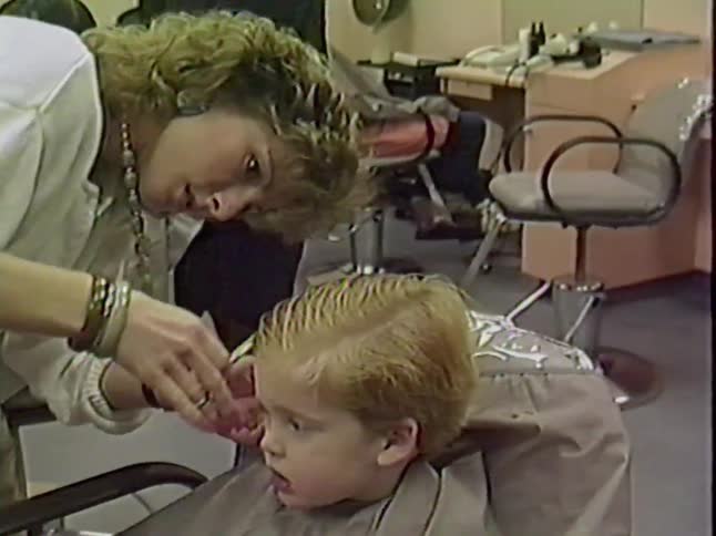 A female hairdresser carefully cuts a toddler's blonde hair with a comb and scissors in a bustling hair salon