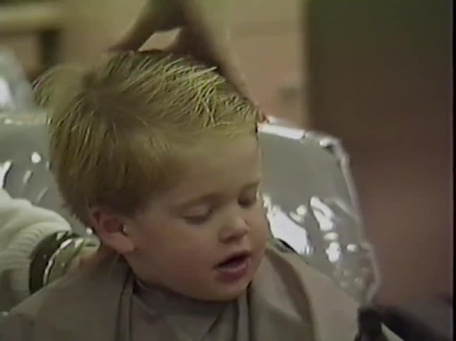 A cute blond toddler boy sits happily in a salon chair while getting his first haircut in this vintage home video