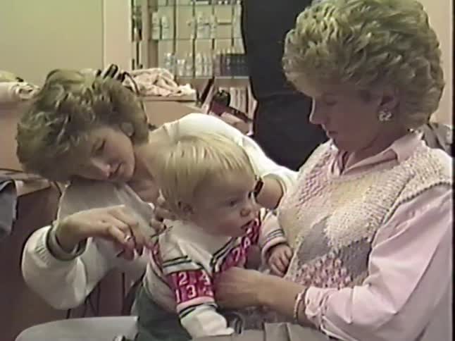 Vintage 1980s footage of a toddler getting his first haircut from a stylist while sitting on his mother's lap