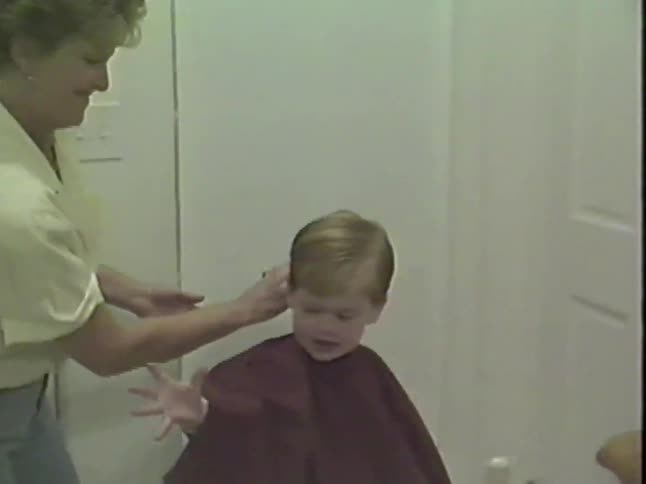 A playful toddler laughs while a woman brushes his face after a haircut, then he takes the brush and playfully dusts her face