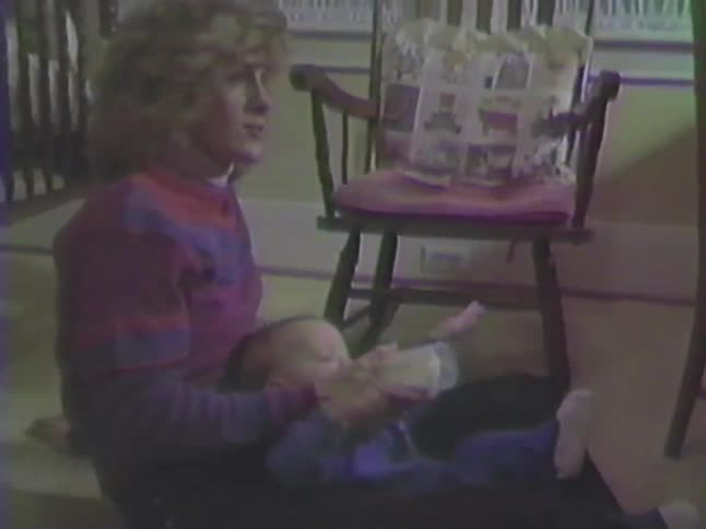 A mother sits on the floor and lovingly feeds her baby a bottle in this vintage home video