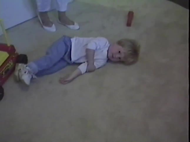 A cute little blonde girl playfully rolls around and lies on a carpeted floor in a vintage home video