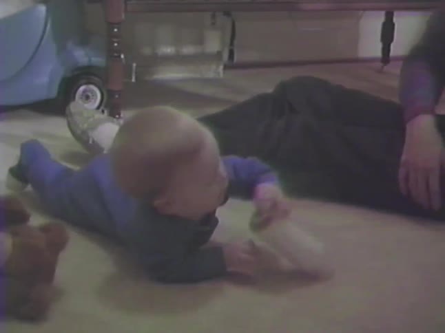 A vintage home movie from the 1990s shows a baby boy in a blue onesie drinking from his bottle on the floor
