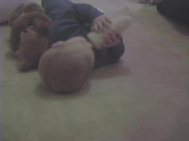 Vintage home video of a baby lying on the floor and drinking milk from a bottle next to a teddy bear