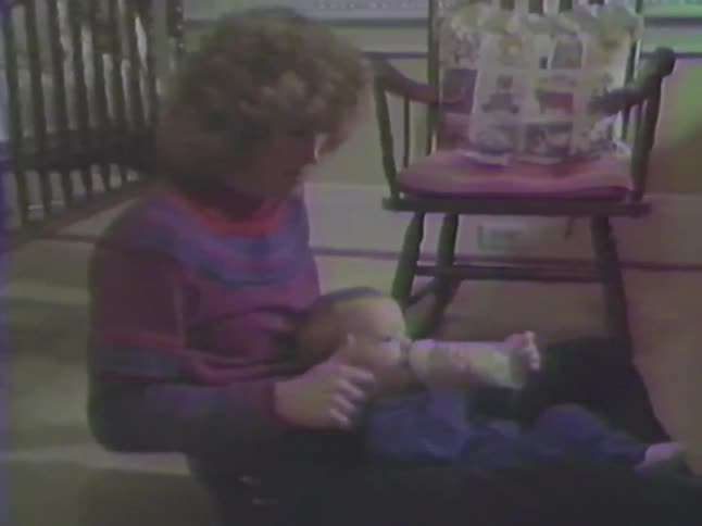 Vintage VHS footage shows a loving mother sitting on the floor and feeding her baby with a bottle in their home
