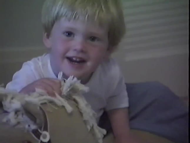 An adorable blonde toddler smiles happily at the camera before the shot zooms out to reveal him playing with his rocking horse