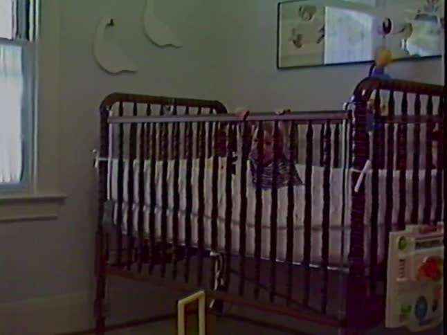 Vintage home video of a baby boy standing happily in his crib before a zoom out reveals his twin in another crib