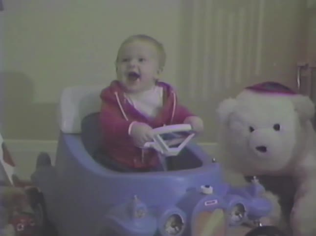A happy baby laughs with excitement while sitting in a blue toy car in a vintage home video