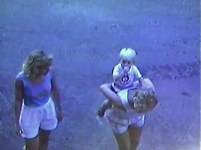 A vintage, high-angle home video captures two women playing with a toddler, giving a piggyback ride and lifting the child in the air