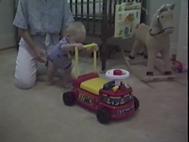 A mother helps her baby learn to walk by holding onto a toy push car in a nursery filled with toys