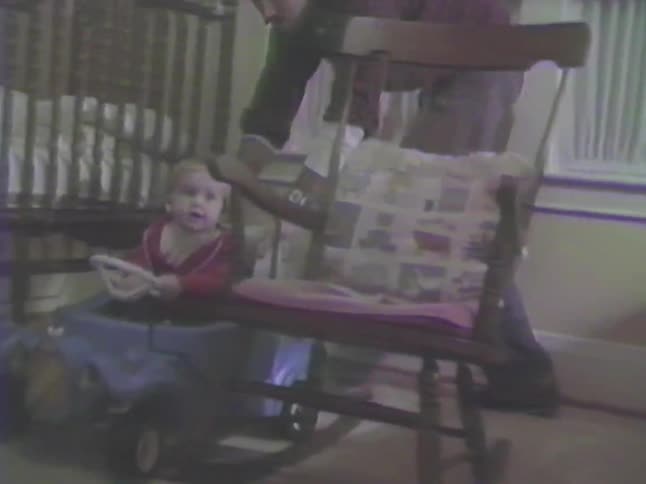 Vintage home video captures a heartwarming moment as a father pushes his smiling toddler in a toy car around the room