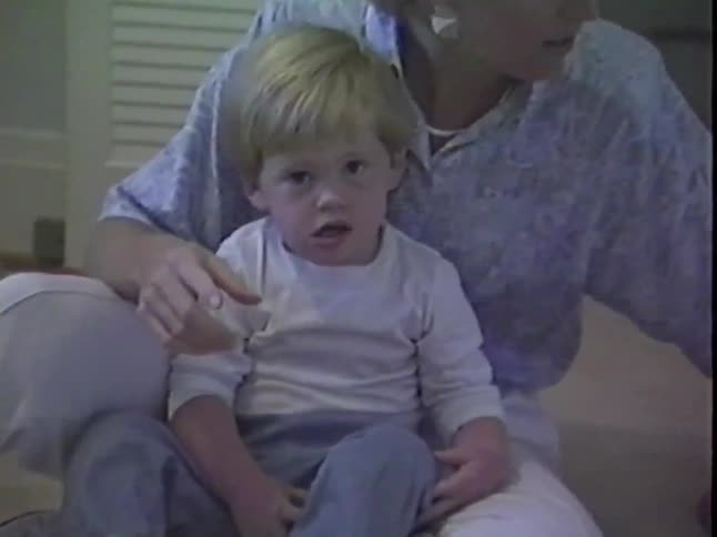 A mother and her young son sit on the floor of a playroom and interact with a teddy bear in this nostalgic home video