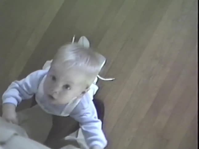 A point-of-view shot of a curious baby with blonde hair and big eyes looking up at the camera while kneeling on a hardwood floor