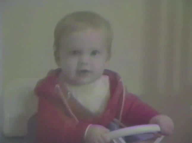 A cute baby in a red hoodie sits in a purple toy car and babbles excitedly in this vintage home video