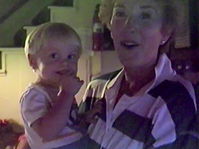 A grandmother holds her cute toddler grandson who sucks his thumb before smiling and pointing at the camera in a nostalgic home video