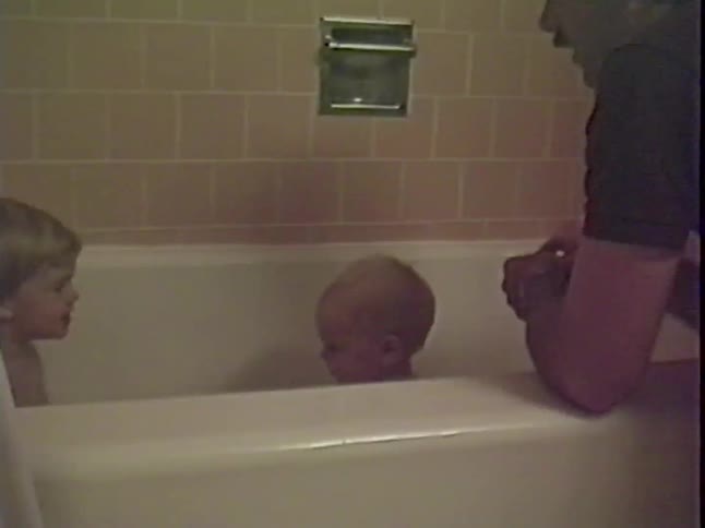 A father watches over his two young children, a baby and a toddler, as they play together in the bathtub