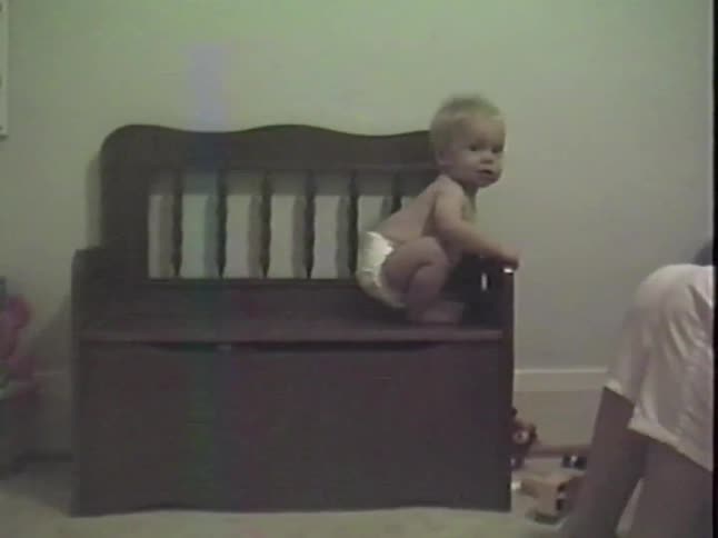 A cute baby in a diaper sits on a wooden toy box and smiles at the camera in a vintage home video