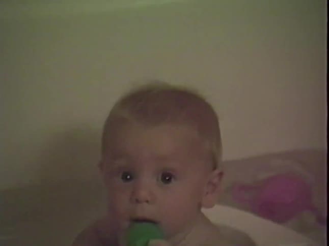 An adorable wide-eyed baby takes a bath, chewing on a toy and looking curiously at the camera