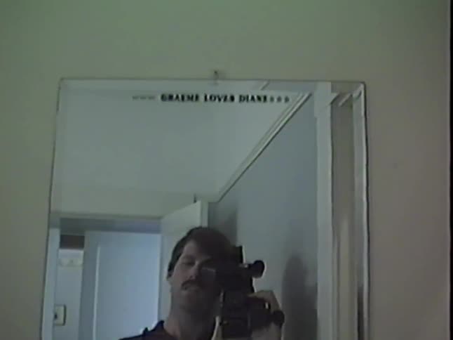 A man with a mustache smiles and waves while filming his reflection in a mirror with a vintage camcorder