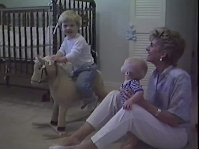 Vintage home video of a happy toddler riding a rocking horse while his mother holds his baby brother in the nursery