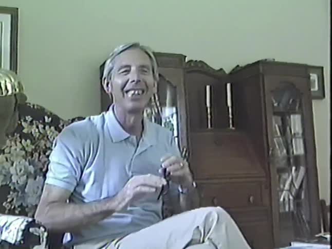 A happy older man with grey hair and glasses sits in a chair and talks animatedly for a home video